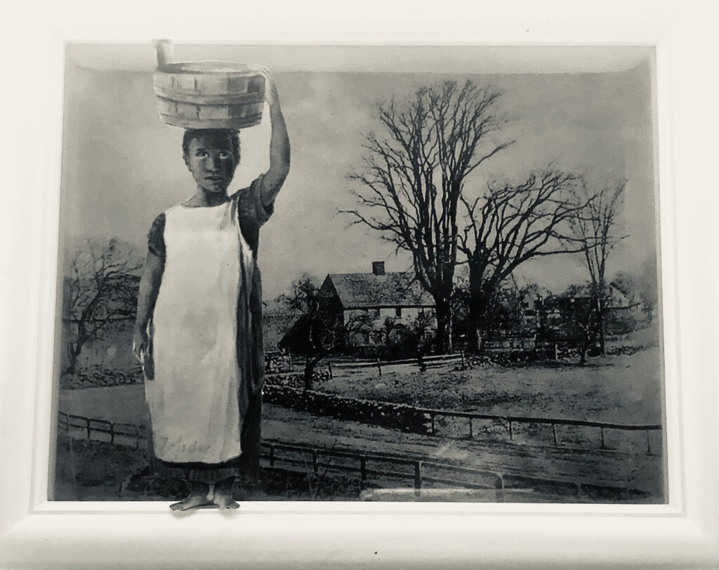 Black girl in white apron with basket on head in front of a building and trees