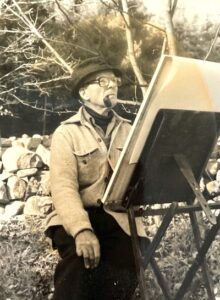 Man in jacket and hat sitting in front of a painting easel outdoors