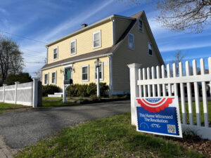 Sing in front of a white picket fence with a colonial house in the background